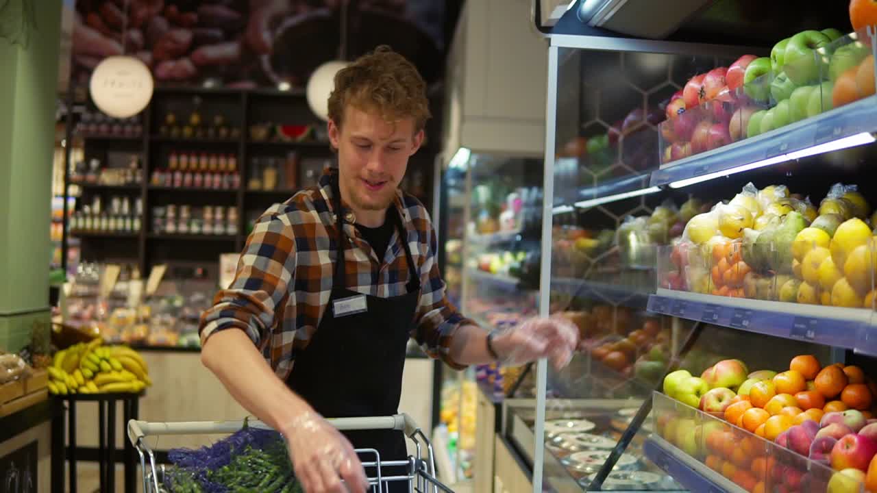 en el supermercado: guapo empleado de almacén con un delantal negro, organizando frutas y verduras orgánicas. agregando manzanas rojas frescas en el estante de la tienda. cámara lenta