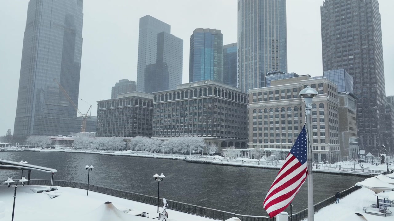 American flag waving over snowy Jersey City waterfront with Manhattan skyline in background during winter snowfall. Urban riverfront scene with patriotic mood and cold cityscape vibes. aerial orbit.