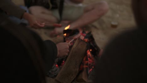 Group-of-young-friends-sitting-by-the-fire-on-the-beach,-grilling-sausages-and-playing-guitar.-Slow-Motion-shot