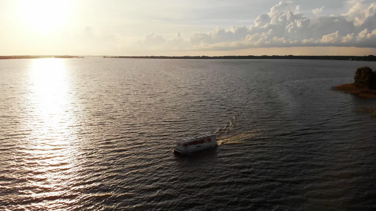 Ampia inquadratura aerea con drone di una barca che naviga in acqua, sole splendente, molo del porto turistico con faro nella baia e palme.