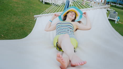 a young girl in a cool bright hat is enjoying the rest on a white hammock in a seaside resort