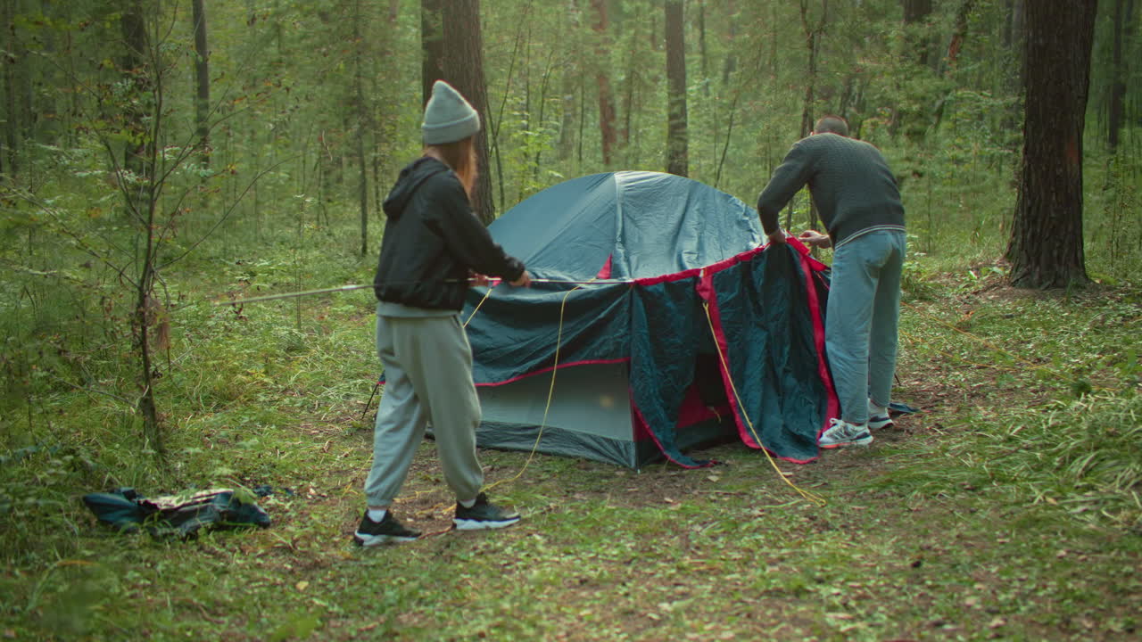 Woman assists boy fixing flexible pole into tent cover in forest clearing, surrounded by trees and grass, collaborating on outdoor camping setup under soft daylight, wearing casual jackets and beanie