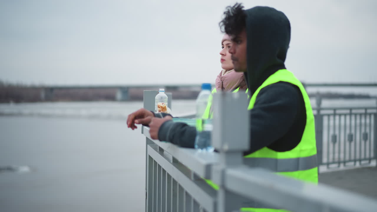 Man in reflective safety vest and hooded sweatshirt eats food from container while leaning on riverside railing in cold weather, woman in pink hat partially visible in background, overcast sky