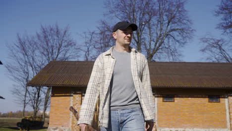 Front-view-of-a-caucasian-man-in-plaid-shirt-and-cap-holding-an-ax-and-walking-outside-a-country-house