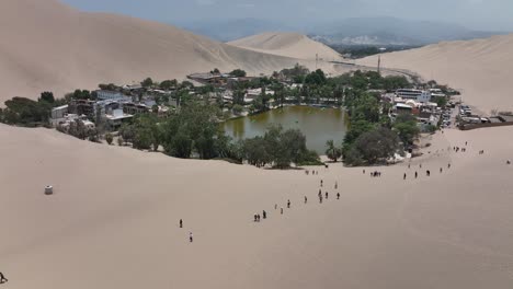 Oasis-Del-Desierto-Huacachina,-Perú-Con-Lago-Y-Palmeras,-Con-Grandes-Dunas-De-Arena-En-El-Fondo