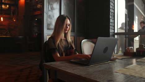 Side-view.-Young-business-woman-sitting-at-table-and-taking-notes-in-notebook.On-table-is-laptop,-smartphone-and-cup-of-coffee.On-computer-screen-graphics-and-charts.-Student-learning-online.-Blogger.