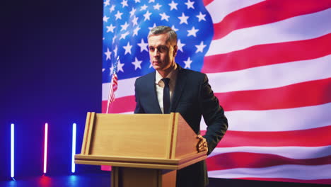 presidential candidate giving speech against usa flag zoom in view of convincing middle aged man in suit standing on podium and speaking against usa flag during election debate