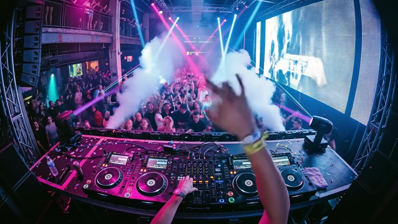 Professional DJ performing at a crowded nightclub with smoke machines and colorful stage lighting