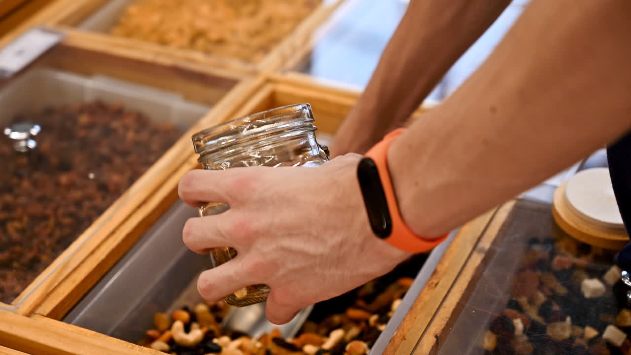 Man's hands filling a glass jar with nuts, dried fruits and berries in supermarket. Ecology idea