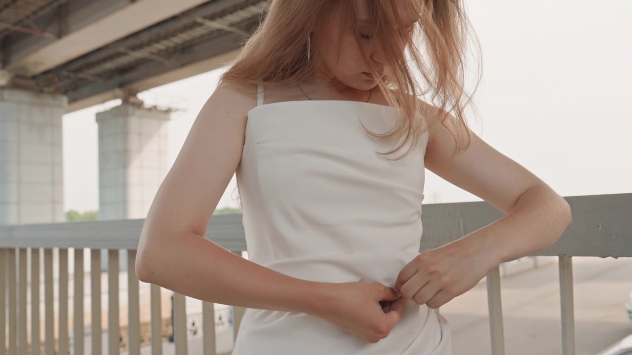 Una donna si misura la vita con un metro a nastro sotto la luce del giorno su un ponte all'aperto. La donna sistema il tessuto del suo vestito bianco e fissa il metro a nastro intorno al busto, concentrandosi intensamente.