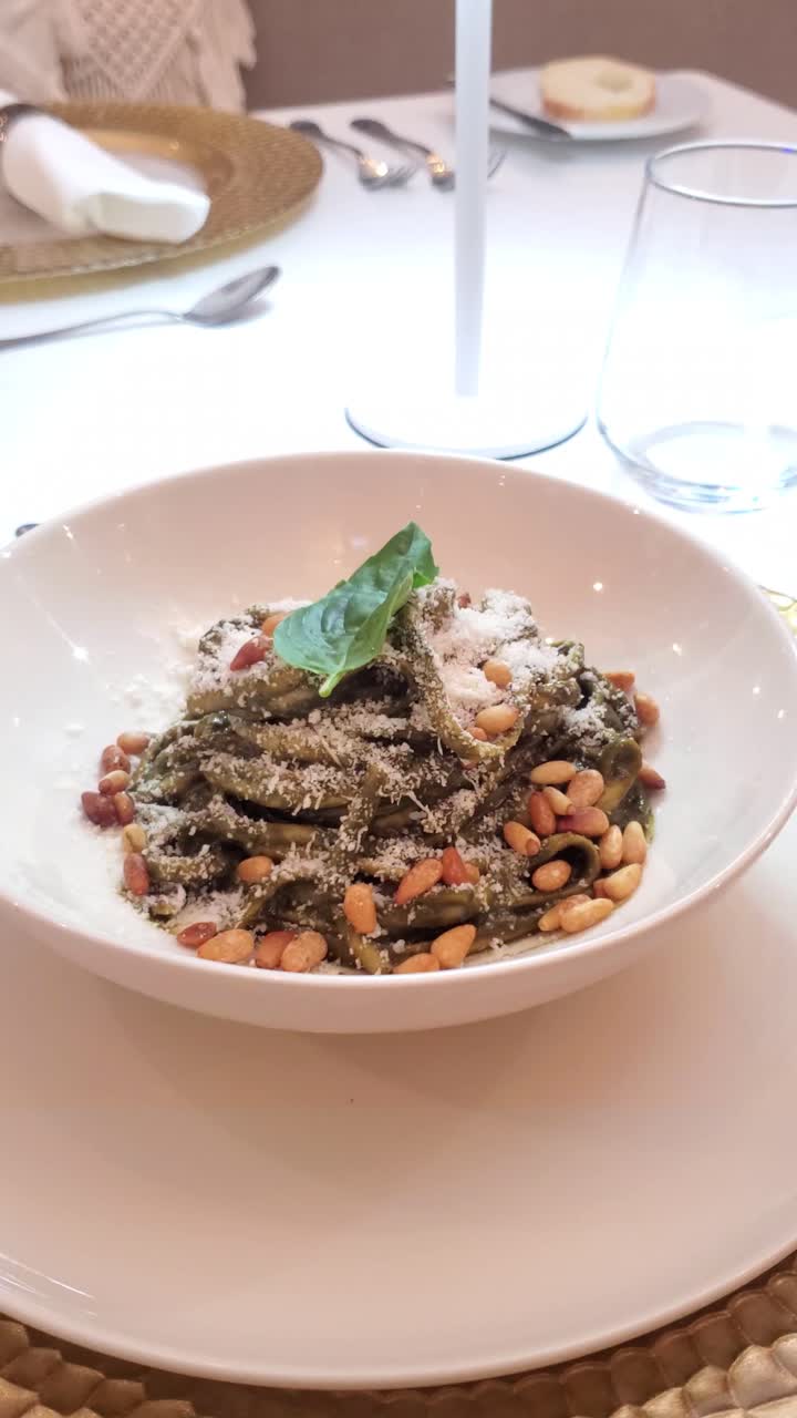 Bowl of green pesto pasta topped with pine nuts and cheese, served on a white plate in restaurant setting