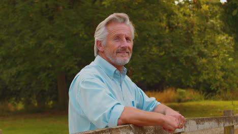 Portrait-Of-Smiling-Casually-Dressed-Mature-Or-Senior-Man-Leaning-On-Fence-On-Walk-In-Countryside