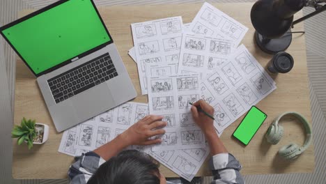 top view zoom out shot of asian male artist drawing storyboard for the film on the table with green screen smartphone and laptop in the studio