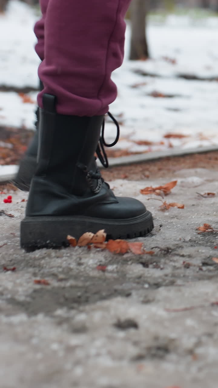 vista trasera de una mujer con botas negras y pantalones de color marrón caminando por un camino cubierto de nieve esparcido de hojas secas, ambiente de invierno helado con una mezcla de suelo helado