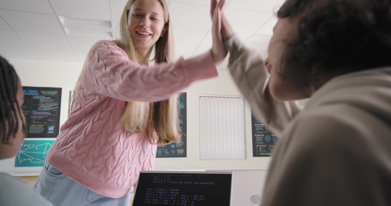 Adolescenti di diverse etnie che seguono un adolescente in piedi con un maglione rosa che indica il codice su un laptop, esultando per una correzione in laboratorio.