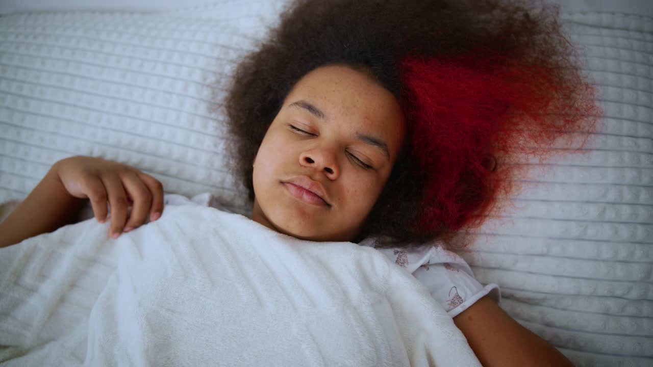 Young African American girl with partially red afro hairstyle lying peacefully, covered by white blanket, resting comfortably in bed with eyes closed during quiet nighttime moment
