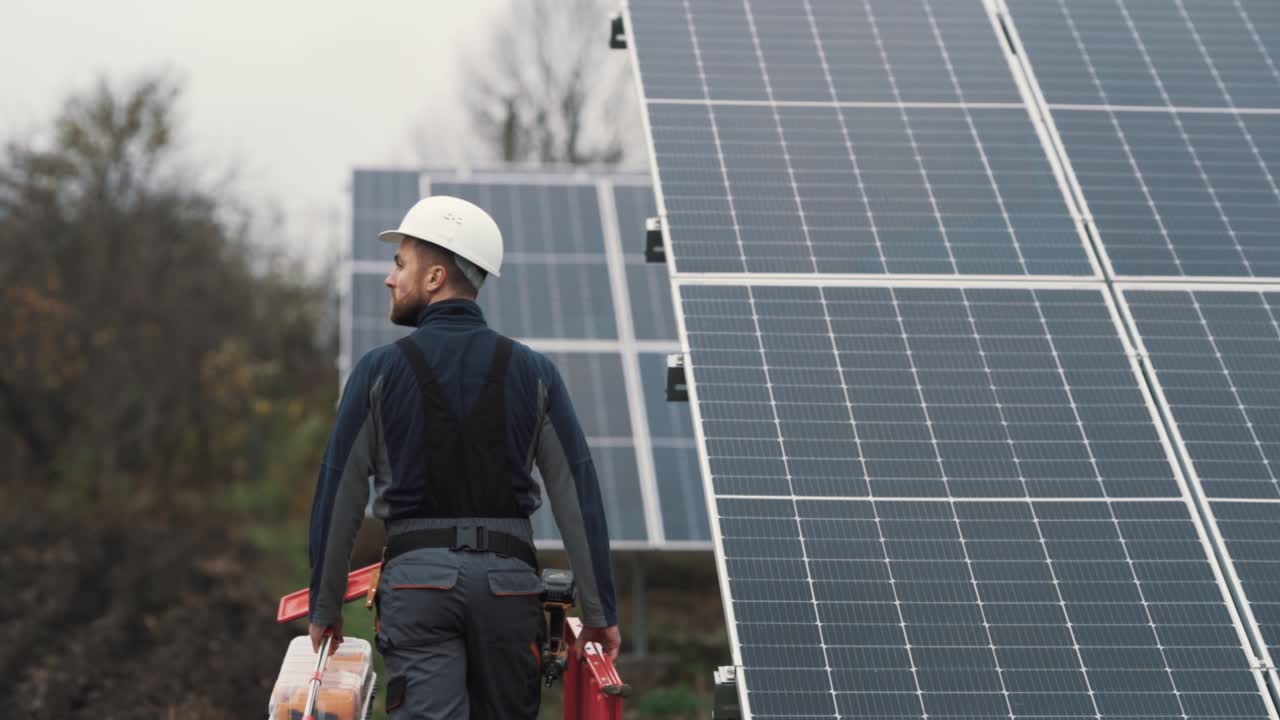Rear view of young man in work uniform with tools in hand inspecting solar panels