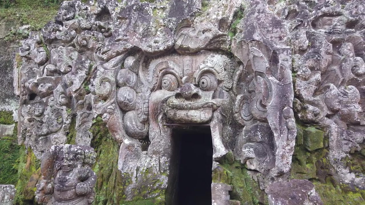 La telecamera panoramica a destra traccia il volto di demone intagliato e ornato all'ingresso di Goa Gajah, catturando l'artigianato balinese in pietra secolare ricoperto di muschio.