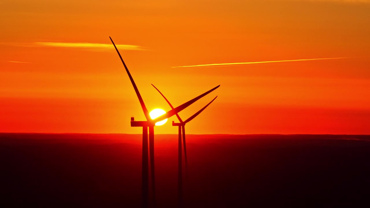 Nahaufnahme einer Windkraftanlage mit der Sonne, die hinter den Rotorblättern am Horizont untergeht.