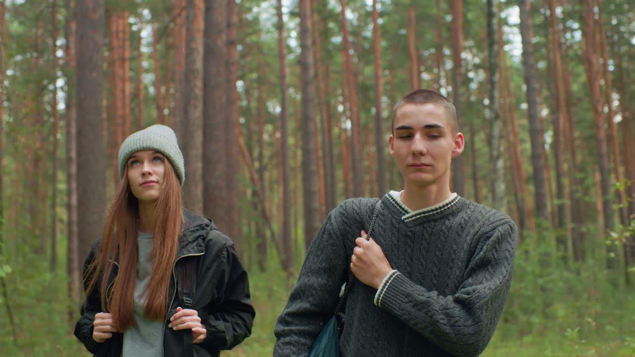Researchers walking through forest trail while boy gestures to show girl something of interest, both dressed casually with backpacks, surrounded by tall trees and greenery