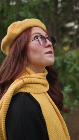 mujer elegante con el cabello rojo largo y los labios pintados, con una boina amarilla y una bufanda, pareciendo feliz mientras mira algo en el fondo, gafas de sol añadiendo a su apariencia chic en un parque exuberante