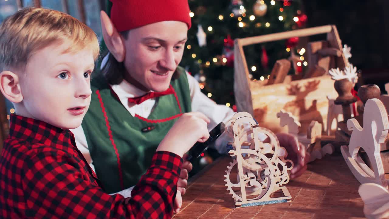 A little boy in a red shirt at the table is played by a wooden toy with gears. Room with a Christmas tree