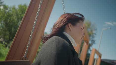 close-up of lady swinging outdoors, hair gently swaying with breeze under bright sunny sky, set against blurred people and greenery, showcasing tranquility, joy, and leisure in a lively park setting