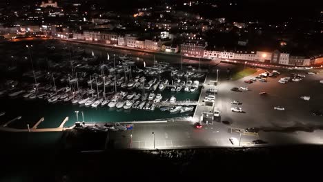 Vuelo-Corto-Y-Elevado-Con-Drones-Nocturnos-Sobre-El-Puerto-Deportivo-Qe-Ii,-El-Puerto-De-San-Pedro,-Guernsey-Con-Grada,-La-Explanada-Glategny-Y-Los-Barcos-En-Sus-Amarres-Y-Las-Luces-Parpadeantes-De-La-Ciudad-Como-Telón-De-Fondo