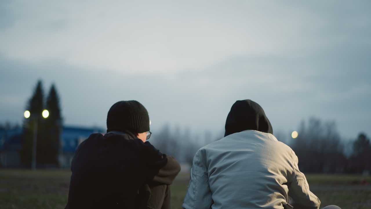 두 소년은 저녁에 잔디에 앉아 있으며, 한 명은 축구공에 왼손을 고 있으며, 빛의 반사와 배경에 빛의 흐릿한 시야와 건물이 있습니다.