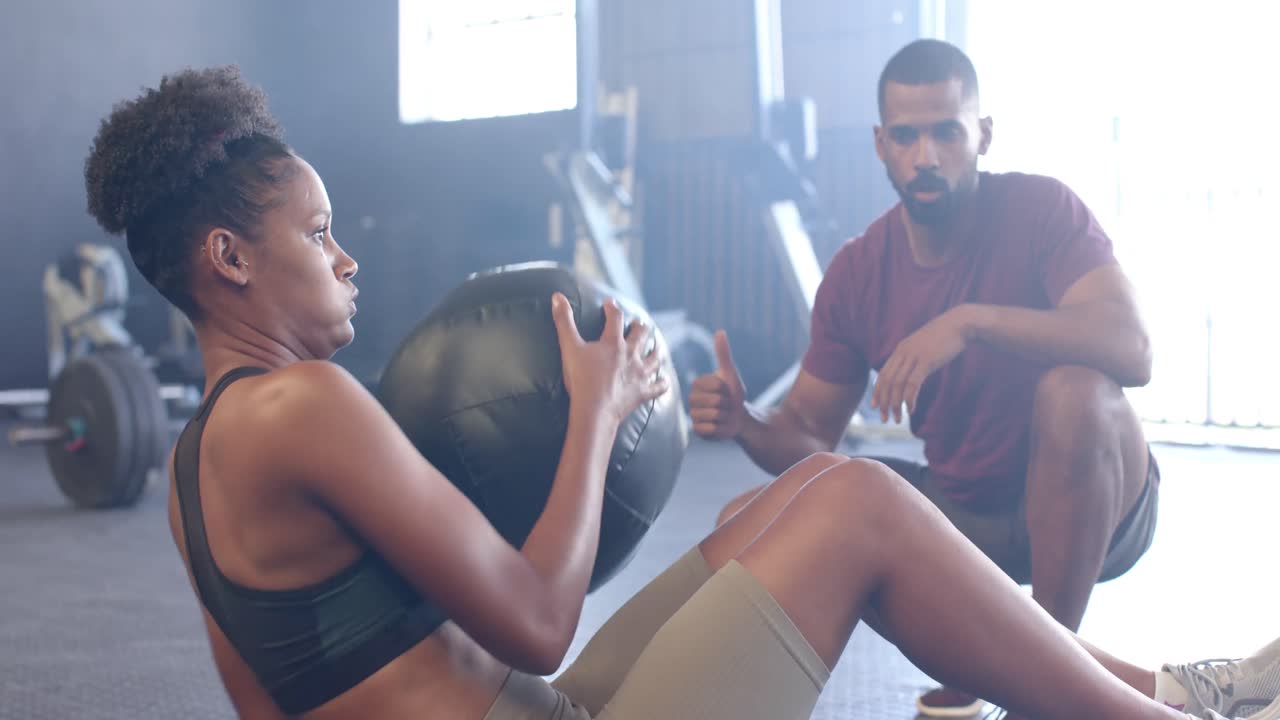 Diverser männlicher Trainer motiviert entschlossene Frau beim Training mit einem Medizinball im Fitnessstudio, in Zeitlupe.