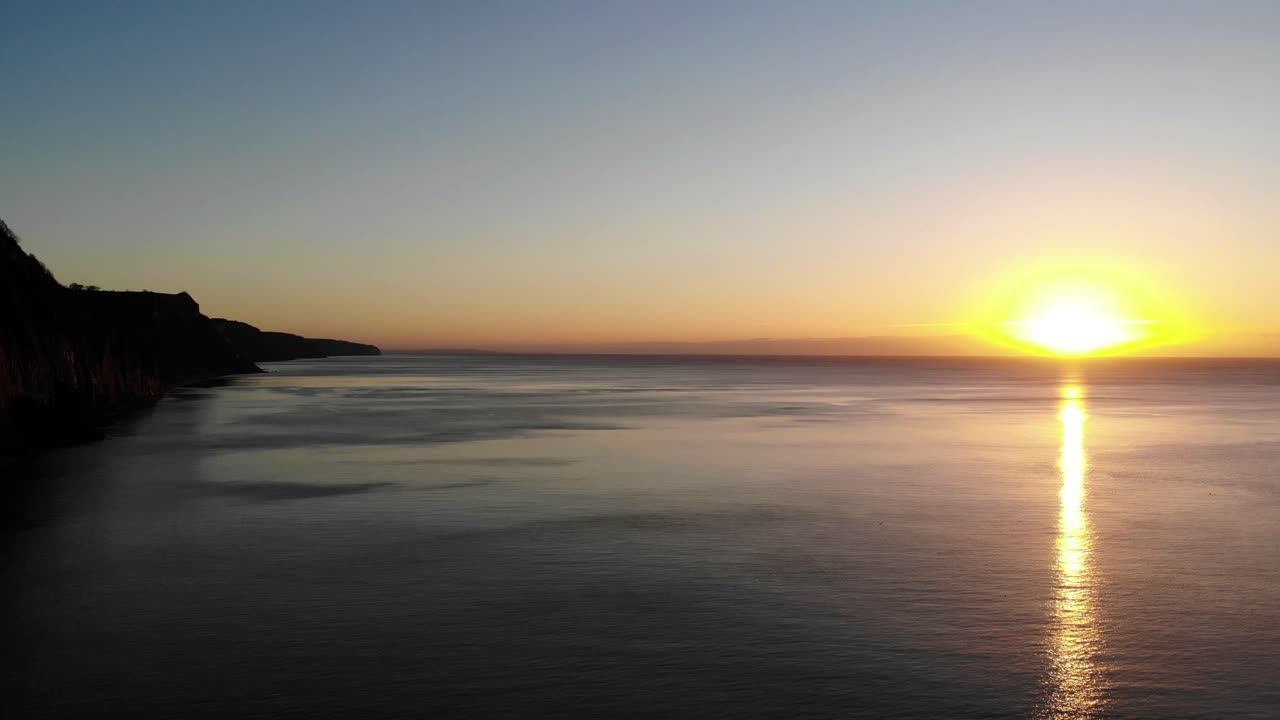Aerial View Of Golden Yellow Morning Sunrise On Horizon Of English Channel With Pan Left Reveal Of Silhouette Of Sidmouth Coastline