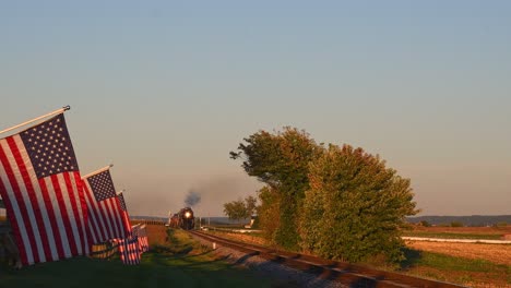 una vista de una línea de banderas americanas que ondean suavemente en una valla junto a tierras de cultivo mientras un tren de pasajeros de vapor que sopla humo se acerca durante la hora dorada