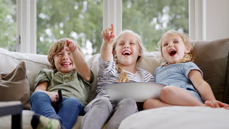 three children sitting on sofa at home laughing and watching tv with popcorn