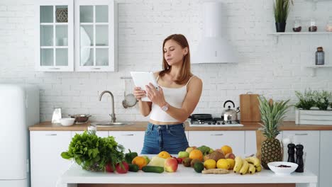 a girl with a tablet in her hands is looking for recipes for cooking healthy food. white kitchen with fruits and vegetables on the table