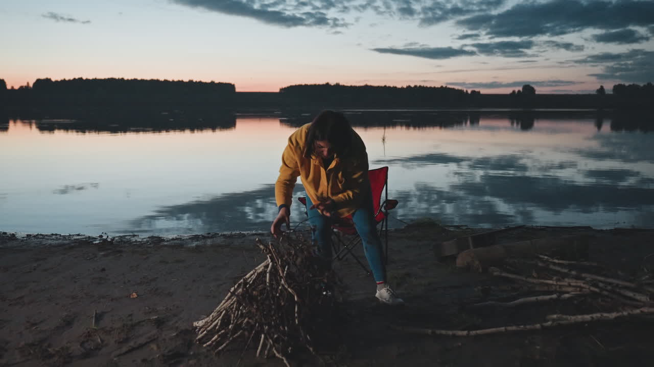 Al crepuscolo, donna in campeggio in riva al lago accatasta legna da ardere accanto a un lago calmo e riflettente al tramonto. Giacca gialla e sedia rossa visibili, raccoglie rami e legna da spiaggia per il falò, riva tranquilla e morbida luce del crepuscolo.