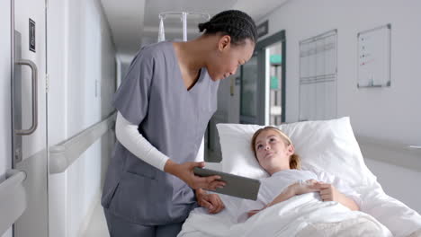 diverse female doctor and girl patient lying on bed using tablet in hospital, slow motion