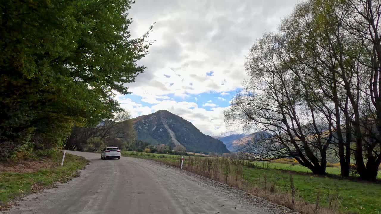 Vehicle travels down rural gravel road, autumn trees, overcast sky, wide angle, steady camera movement