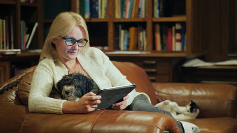 the woman uses the tablet at home she has a puppy in her arms and in the background a cat plays with