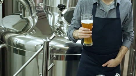 young cheerful brewmaster looking at glass of freshly brewed unfiltered beer and smiling at camera