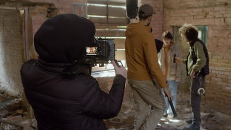 cameraman and team production recording a movie scene with a redheaded girl and man wearing black balaclava in a ruined building