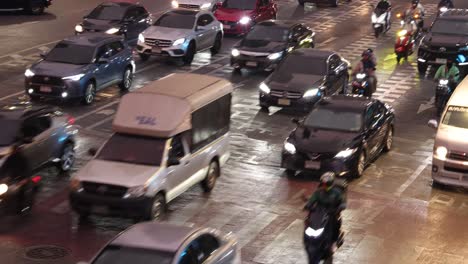 diversos vehículos navegan por una encrucijada húmeda e iluminada de la ciudad