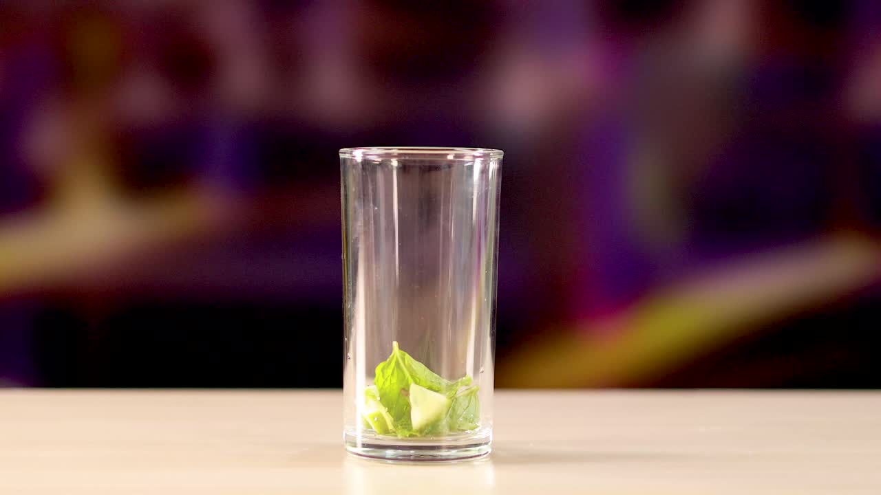 Ice cubes fall into a glass with lime wedges on a table. Bright lighting and a blurred background create a lively atmosphere