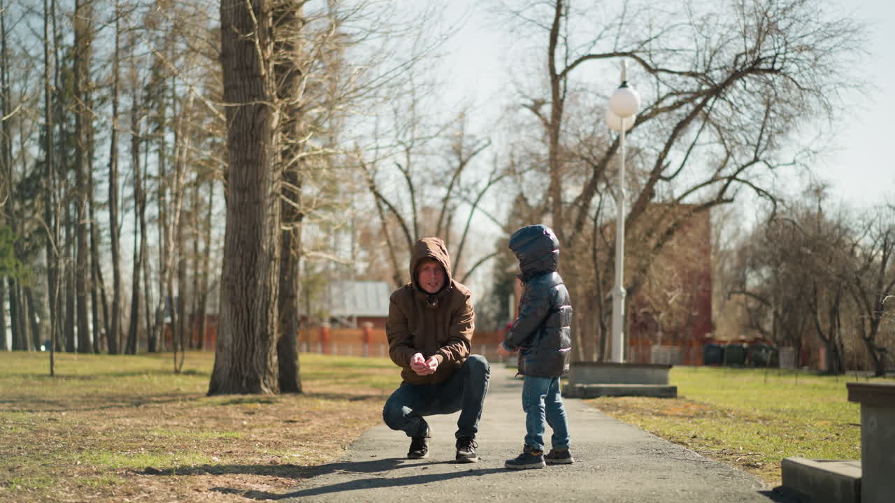 갈색 재을 입은 아버지가 공원 길에서 혀서 공중에 입자를 방출하는 동안 그의 어린 아들은 반이는 검은 재과 청바지를 입고 흥분하여 그들을 잡기 위해 달려갑니다.