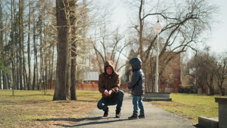a father, dressed in a brown jacket, squats on a park path as he releases some particles into the air, while his young son, wearing a shiny black jacket and jeans,excitedly run to catch them
