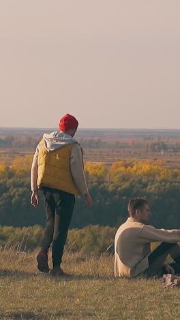 stylish guy in warm vest approaches sad friend sitting at campfire on river bank against dense forest in morning slow motion