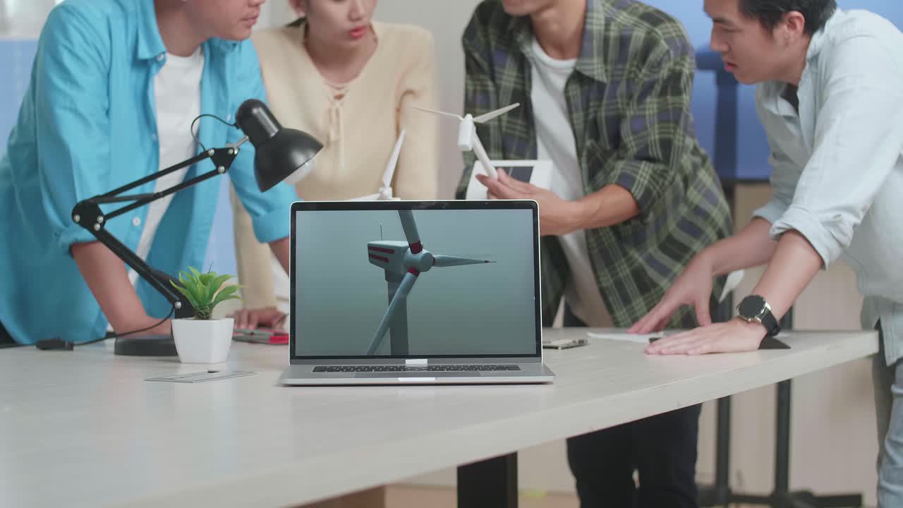una computadora portátil en la mesa está mostrando el video de la turbina eólica mientras un grupo de ingenieros asiáticos discuten sobre el trabajo en la oficina