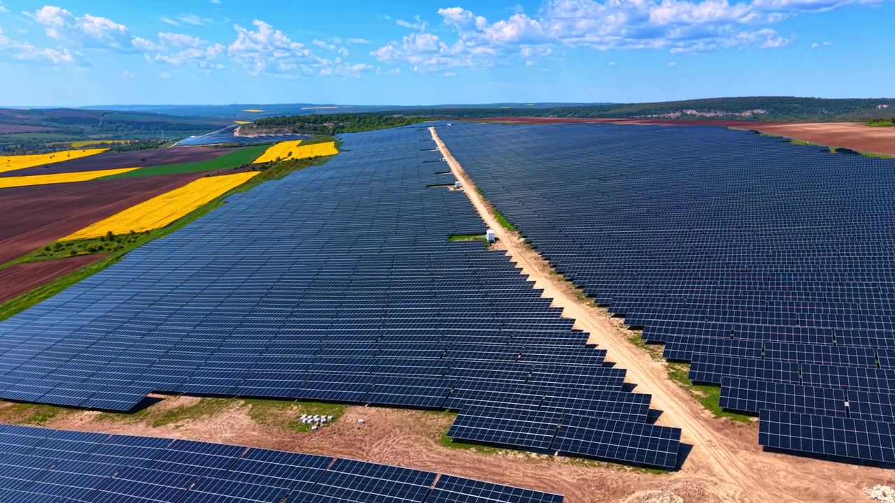 Solcellepark delt af grusvej, hvide udstyrskabinetter står ved siden af fjerntliggende agerland