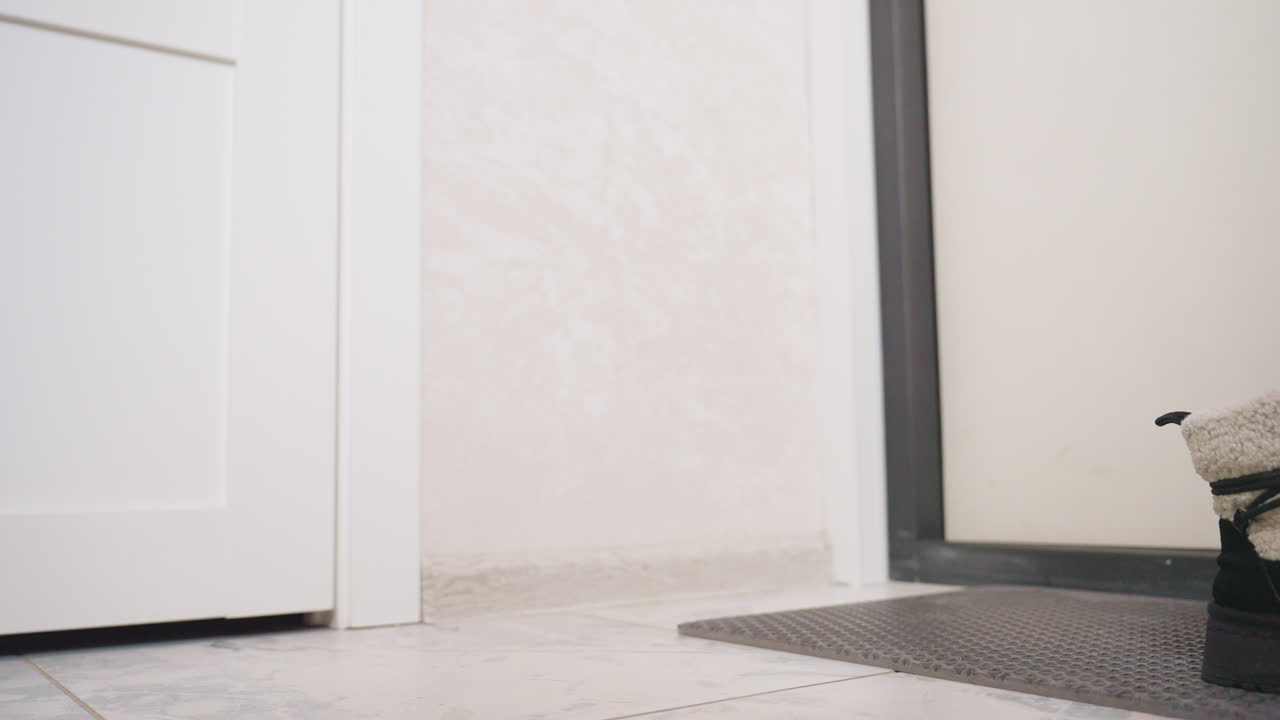 Legs of woman wearing black trousers and white socks walking past wooden shoe rack with slippers and boots in tiled hallway, capturing subtle motion and casual indoor routine