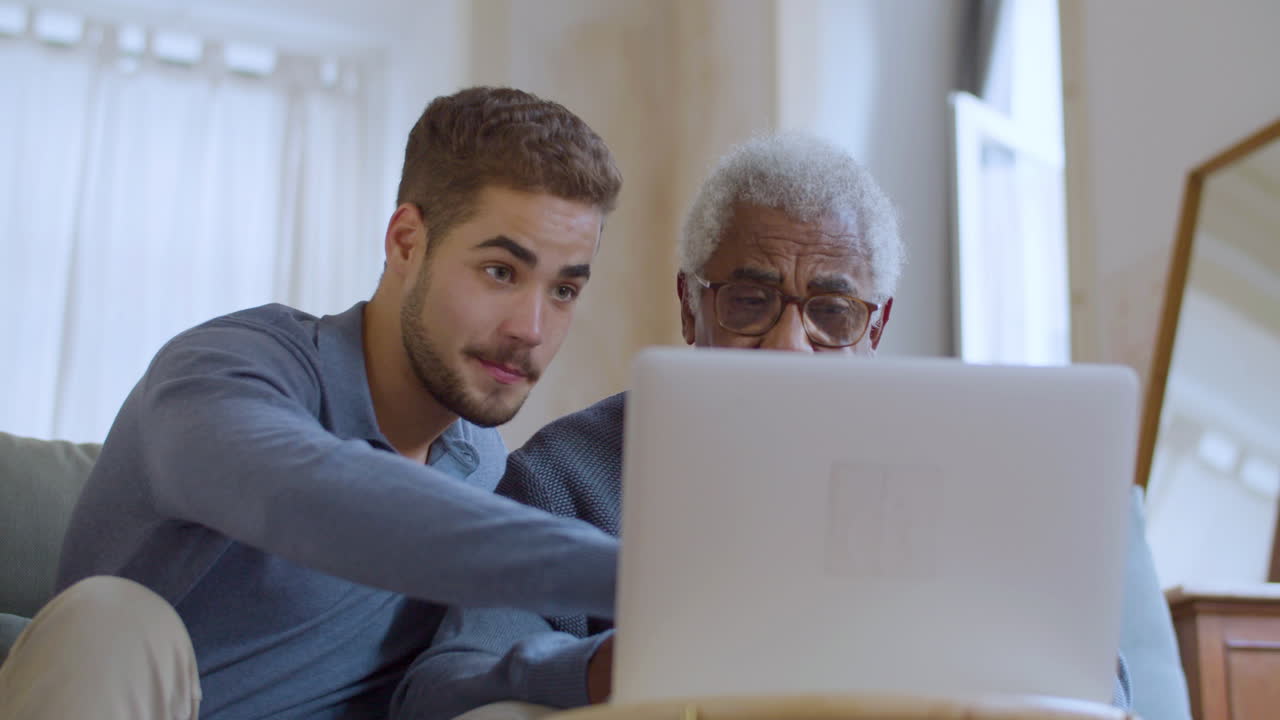 Young Caucasian guy helping senior Black man with using laptop.