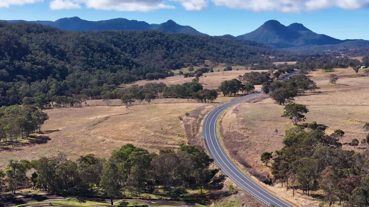 Drone glides above curving road, dry fields, and forested hills under bright daylight, smooth movement